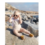 Bebé con sandalias Muris Rio Mini mostaza, peto beige y gafas de sol sentado en una roca junto a la playa